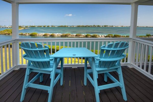 A patio with two blue chairs and a matching table overlooks a serene body of water on a sunny day, with a lush landscape in the background.