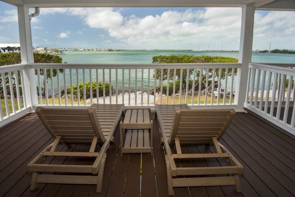 Two lounge chairs and a small table are on a wooden deck, overlooking a serene body of water with trees and a partly cloudy sky in the background.