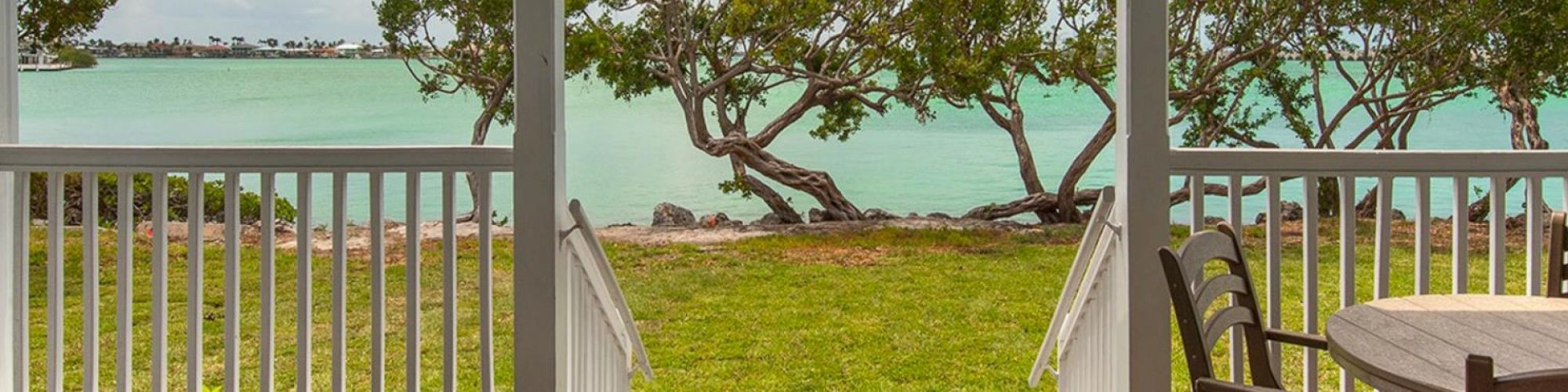 A wooden deck with chairs overlooks a grassy lawn, trees, and a turquoise body of water, framed by two white posts.