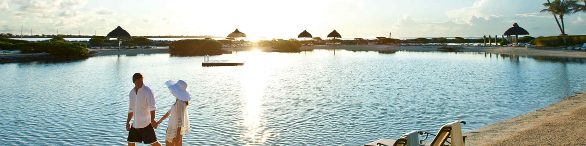 A couple walks along a sandy beach next to a calm body of water at sunrise or sunset, with empty lounge chairs and palm trees in the background.