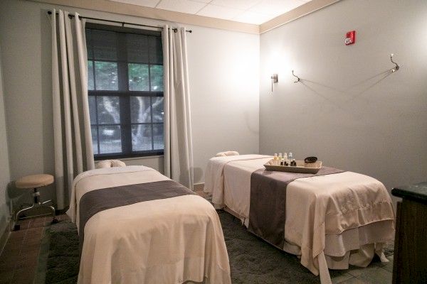 The image shows a serene massage therapy room with two massage tables, a window with curtains, a stool, and a tray of oils on one table.