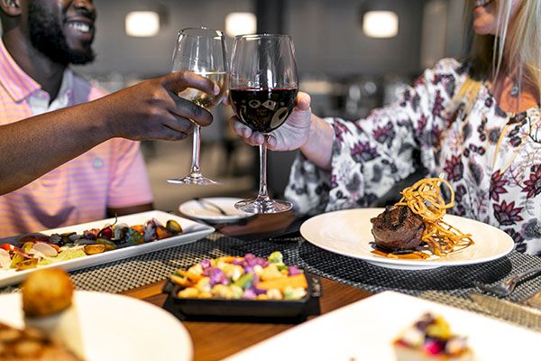 Two people clink wine glasses over a table of gourmet dishes, enjoying a meal together in a restaurant setting.