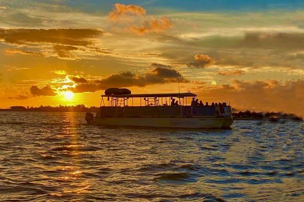 A boat with passengers sails on a body of water during a picturesque sunset with orange and yellow hues in the sky.