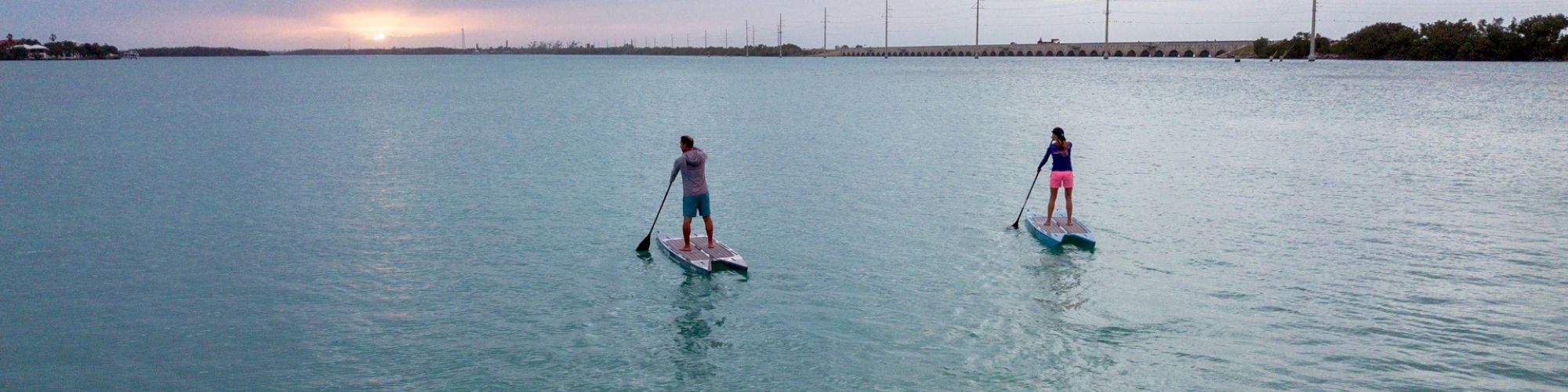 Two people paddleboarding on a calm body of water at sunset, with a long bridge or causeway in the background ending the sentence.