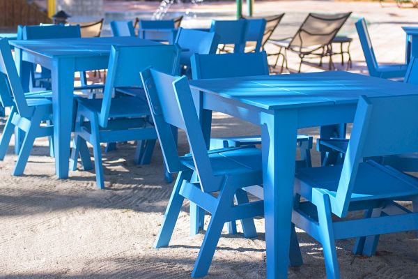 The image shows several blue wooden tables and chairs arranged outdoors on a sandy surface, with some additional seating in the background.