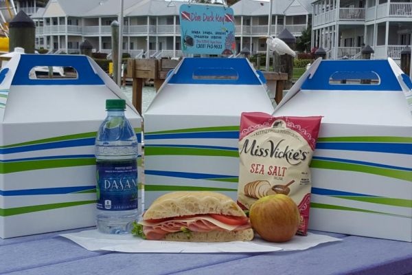 Three boxed lunches, a bottle of water, a bag of chips, a sandwich, and an apple are displayed on a table near a dock.