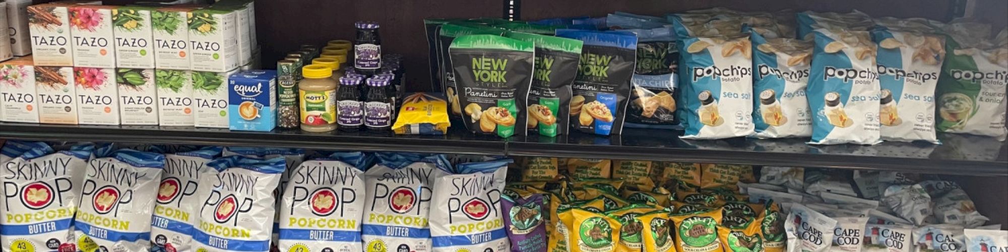 A shelf is stocked with various snacks including popcorn, chips, crackers, tea, and coffee filters, with small bottles of sanitizer on the top right.