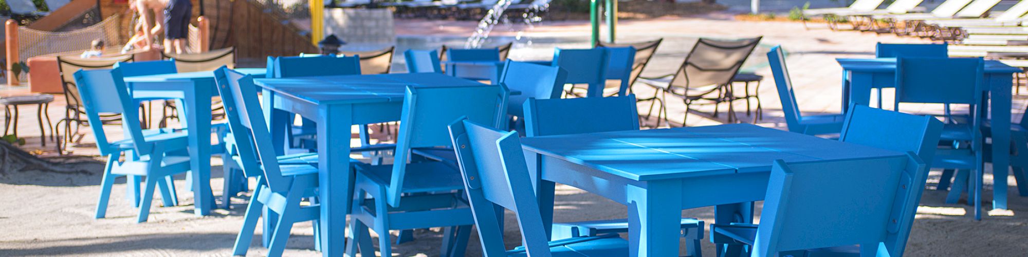 Outdoor seating with blue tables and chairs is set up on sandy ground near lounge chairs and a children's play area in a tropical setting.