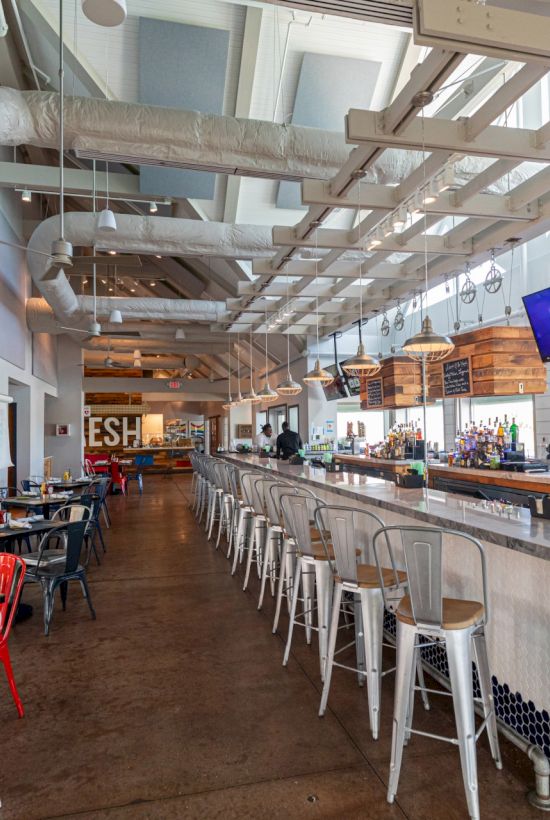 An empty, modern restaurant with an industrial design, featuring a long bar with stools, tables, and screens displaying images.