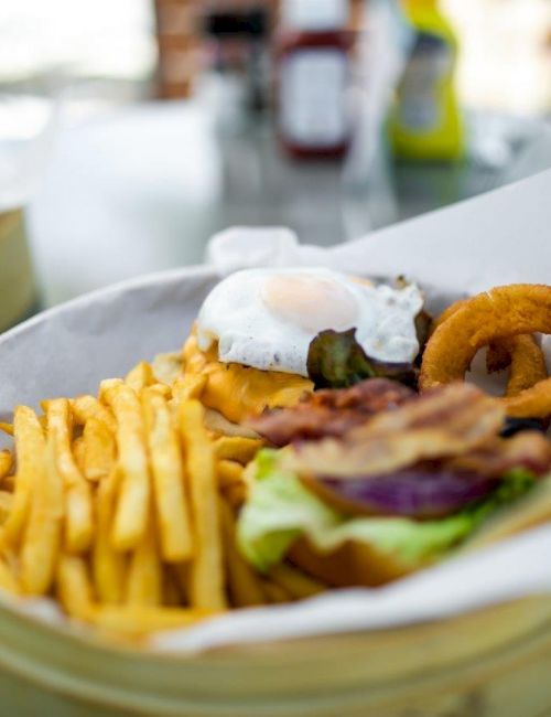 A burger topped with a poached egg, bacon, and an onion ring, served with a side of french fries, sits in a paper-lined basket on a table.