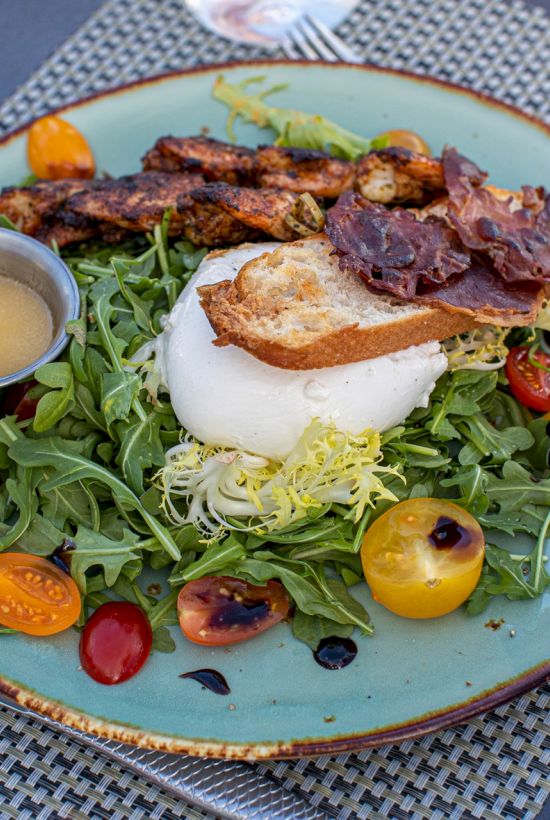 A fresh salad with arugula, cherry tomatoes, grilled chicken, a poached egg, toasted bread, and a side of dressing on a green plate.