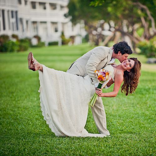 A couple is embracing outdoors, with the man leaning back the woman holding flowers, in a joyful pose on a grassy lawn.