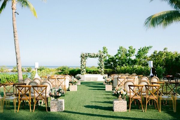 An outdoor wedding setup features an archway with greenery, aligned chairs, and decorative floral arrangements on a grassy area with palm trees.