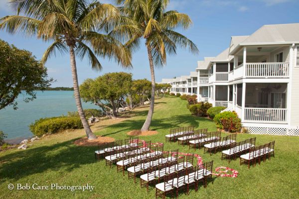 The image shows a wedding setup with rows of chairs on a lawn near the water, palm trees, and a white building with balconies.