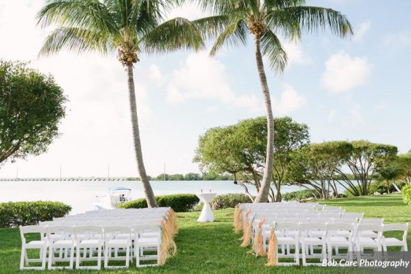 An outdoor wedding setup with rows of white chairs arranged under palm trees, overlooking water; the scene is sunny and serene.