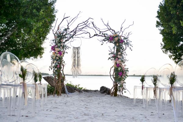 This image shows a beach wedding setup with clear chairs in rows, leading to an arch made of branches and flowers in front of a water backdrop.