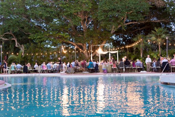 The image shows an outdoor event by a pool, with people seated at tables, string lights overhead, and trees in the background.