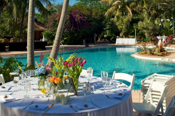 An outdoor setup with a round table by a pool, decorated with flowers and chairs, surrounded by tall palm trees and lush greenery under a clear blue sky.