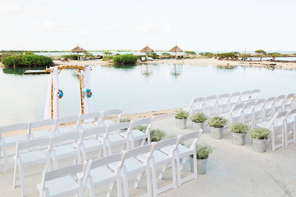 This image shows a beachside wedding setup with rows of white chairs facing a bamboo arch, with plants placed along the aisle and water in the background.