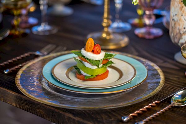 A beautifully plated appetizer with layers of tomato, mozzarella, and basil sits on a decorative plate on a set dining table.