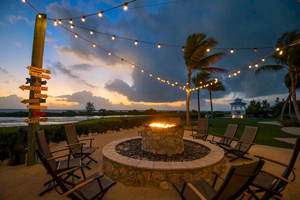 The image shows a scenic evening view with a fire pit surrounded by chairs, string lights, and palm trees beside a shoreline.