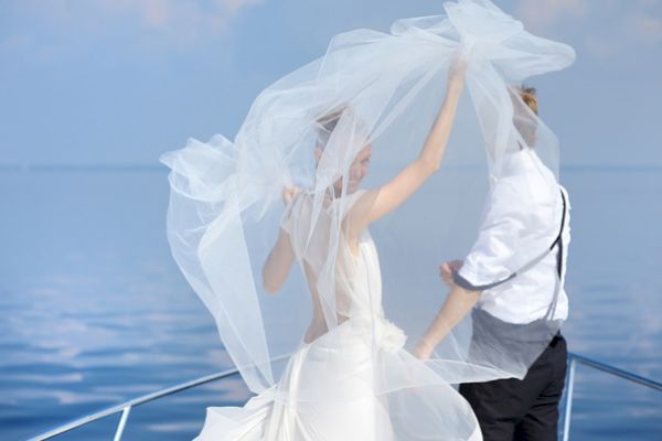 A bride and groom stand on a boat, with the bride's veil billowing in the wind, against a serene ocean backdrop ending the sentence.