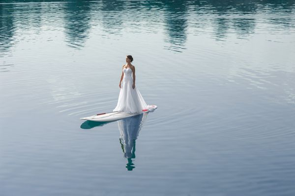 A woman in a white gown is standing on a paddleboard in the middle of a calm, reflective body of water, creating a serene scene.