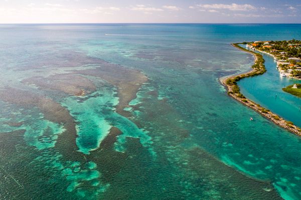 Aerial view of a coast with turquoise waters, coral reefs, and a strip of land with some structures, surrounded by the ocean under a clear sky.