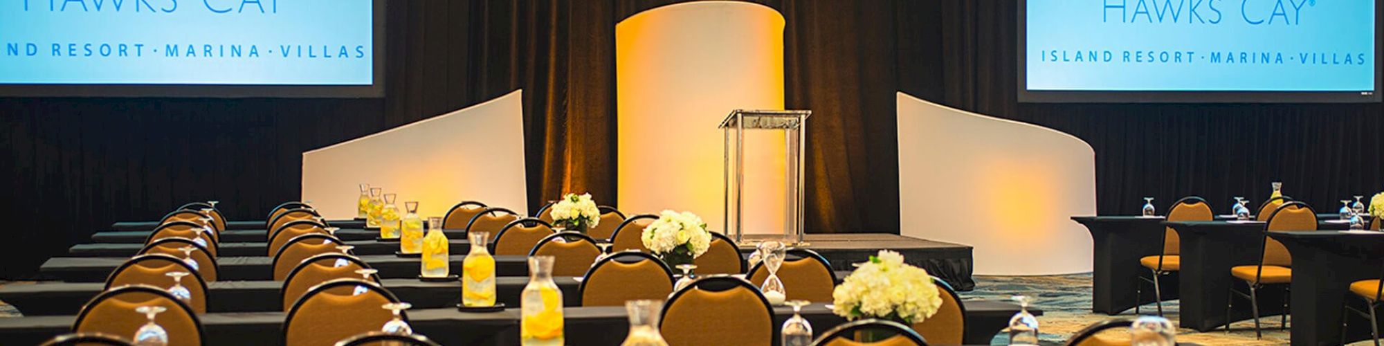 The image shows a conference room setup with rows of tables and chairs, a podium, and two projection screens displaying 