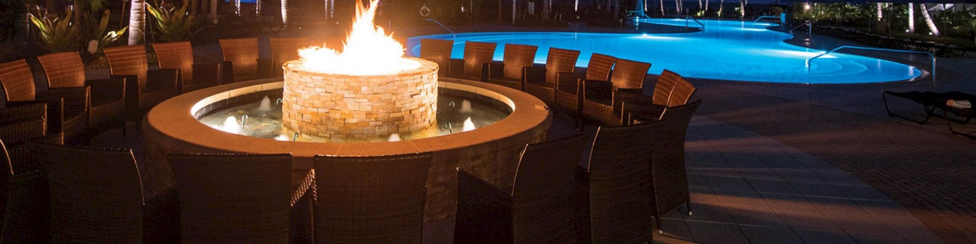 An inviting outdoor seating area with a fire pit, surrounded by wicker chairs near a lit swimming pool, set against a nighttime backdrop.