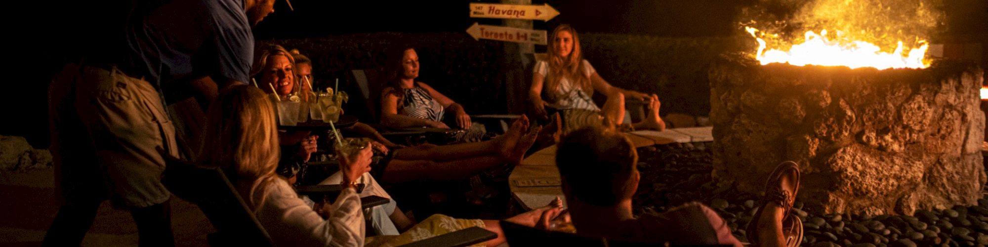 A group of people are sitting around a fire pit at night, enjoying drinks, with directional signs in the background pointing to various places.