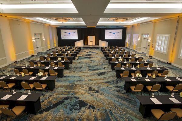 A large conference or meeting room with rows of tables and chairs facing a central stage and two projection screens. The room is well lit.