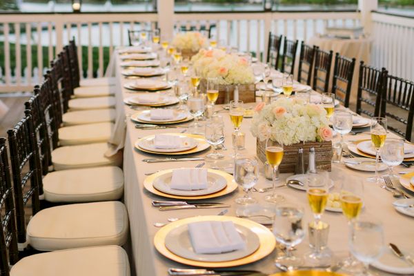 A long, elegantly set dining table under a chandelier on a porch overlooking a scenic body of water, featuring gold accents and neatly arranged settings.