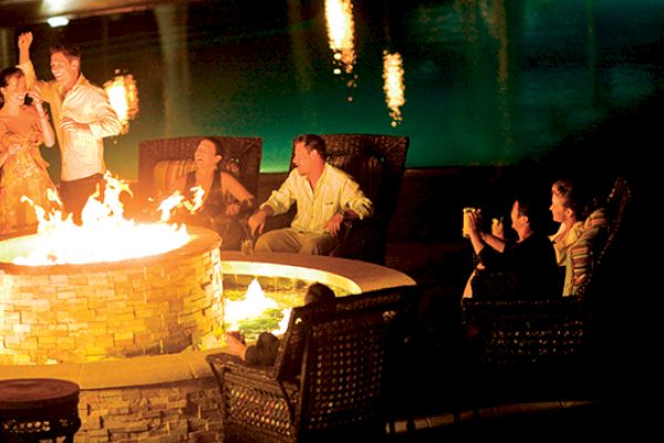 People are gathered around a fire pit, with some standing and others sitting, enjoying a nighttime outdoor setting near a pool.