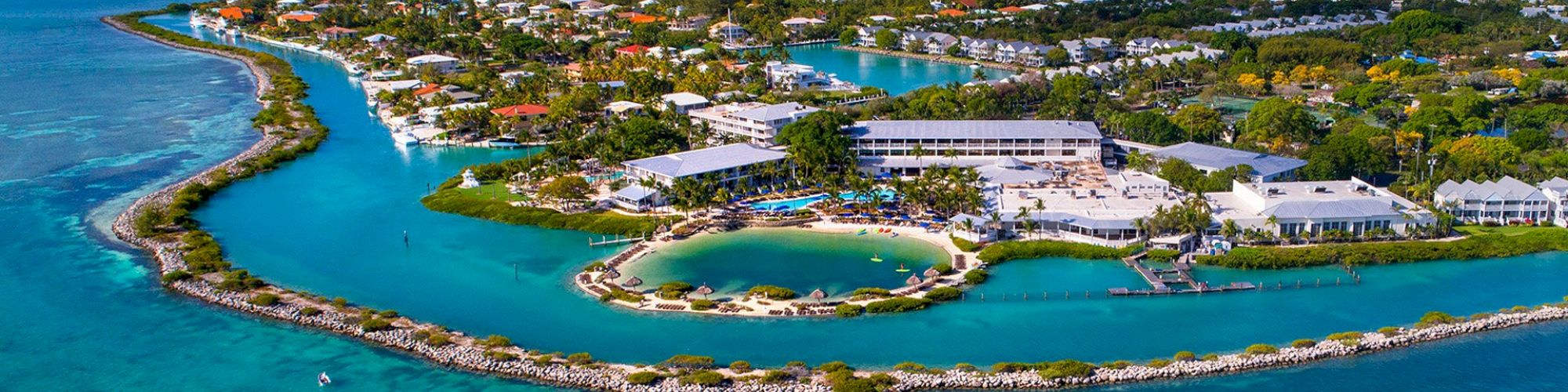 An aerial view of a coastal resort surrounded by turquoise water, featuring numerous buildings, a swimming pool, and lush greenery, forming a scenic landscape.