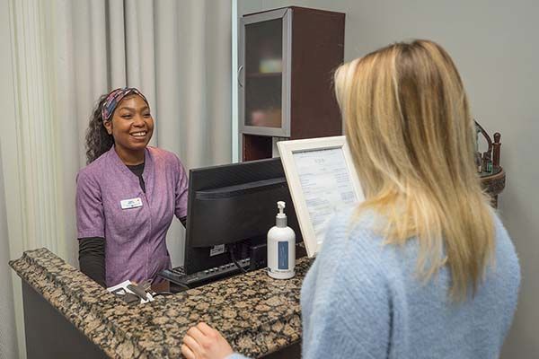 A receptionist in a purple uniform smiles at a blonde woman in a blue sweater standing at a granite counter with a computer and sanitizer.