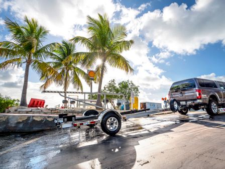 A pickup truck with an attached boat trailer is on a boat ramp by palm trees and a bright blue sky with scattered clouds, capturing a sunny day.