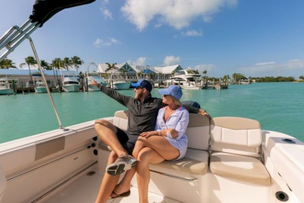 A couple is sitting on a boat with turquoise water and a marina in the background, pointing towards the shore and enjoying the sunny weather.