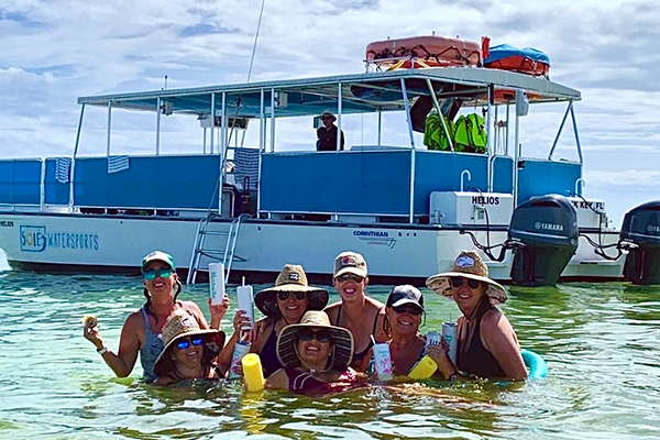 A group of people wearing hats and swimsuits are in the water holding drinks with a boat and its crew behind them under a partly cloudy sky.