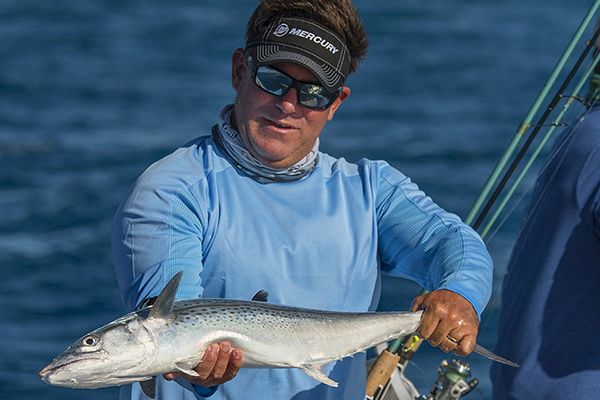 A person wearing sunglasses and a headband holds a freshly caught fish while standing on a boat in the water.