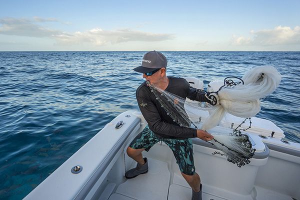 A man on a boat is casting a fishing net into the ocean under a clear blue sky.