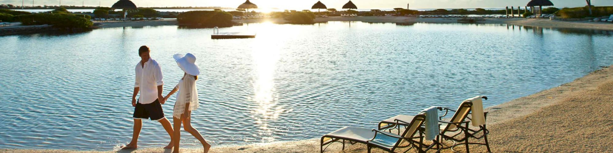 A couple walks hand in hand along the beach at sunset, with lounge chairs in the foreground and thatched umbrellas in the background, on a calm day.