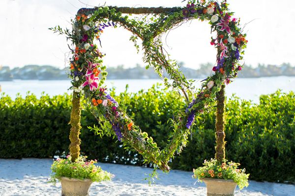 A heart-shaped floral arch made of greenery and flowers stands on the beach with a scenic lake view in the background.