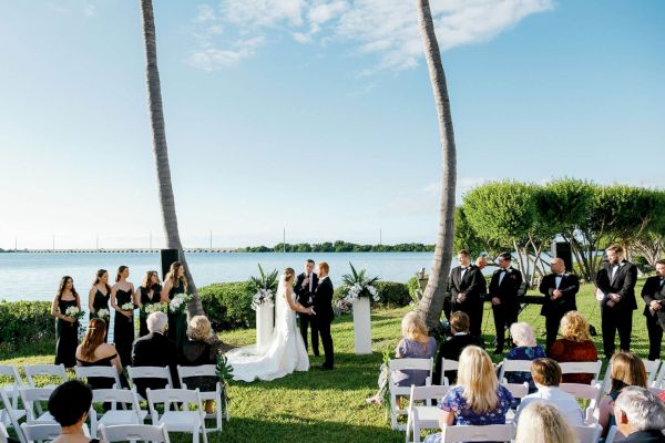 A wedding ceremony is taking place outdoors by a lake, with guests seated on white chairs under palm trees as a couple stands at the altar.