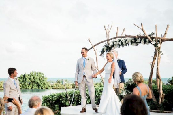 A couple stands at an outdoor wedding ceremony under a rustic wooden arch adorned with flowers, surrounded by guests near a water backdrop.