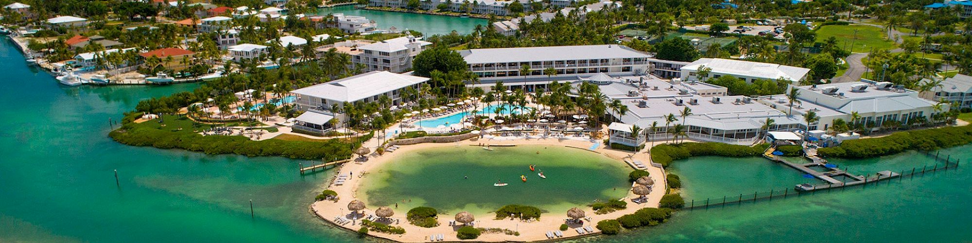 An aerial view of a resort with a private beach, surrounded by turquoise water and lush greenery, featuring multiple buildings and recreational areas.