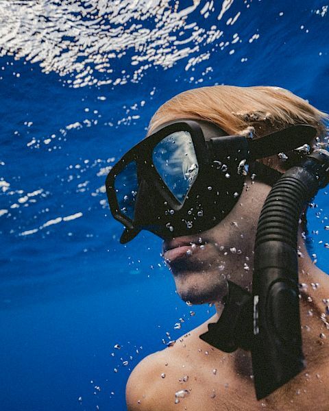 A person is underwater, wearing a snorkel mask and looking upward. Bubbles and reflections are visible in the surrounding blue water.