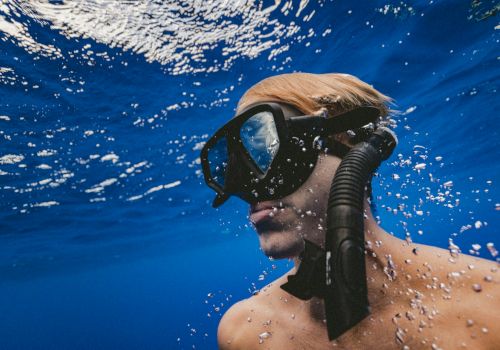 A person is underwater, wearing a snorkel mask and looking upward. Bubbles and reflections are visible in the surrounding blue water.