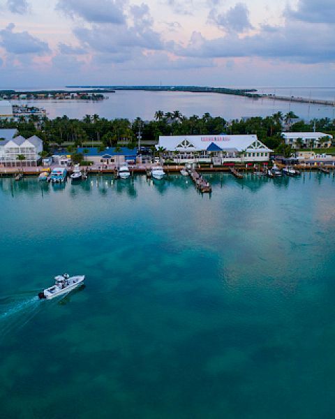 An aerial view captures a tranquil coastal town with buildings, docks, and a boat gliding across calm, clear waters under a partly cloudy sky.