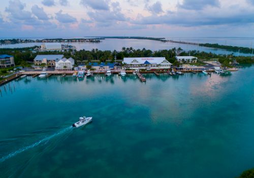 An aerial view captures a tranquil coastal town with buildings, docks, and a boat gliding across calm, clear waters under a partly cloudy sky.